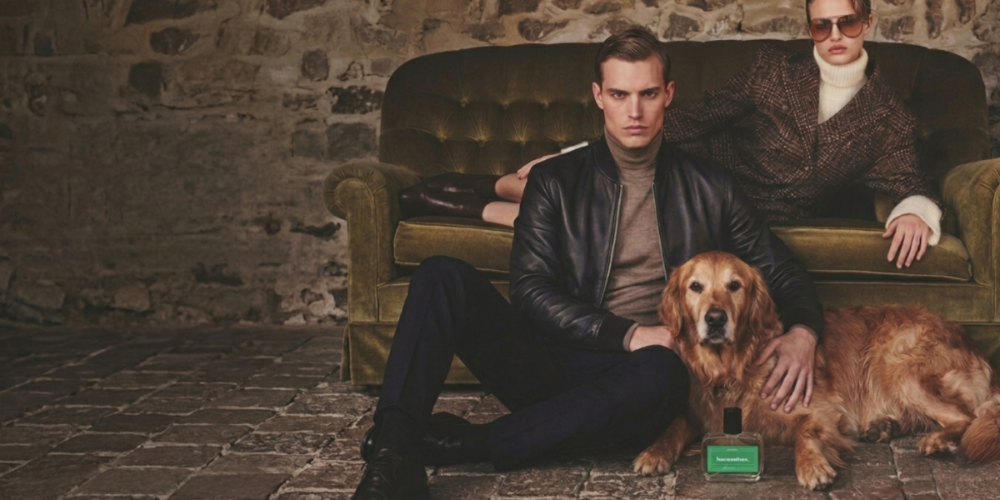 A lifestyle fashion photograph featuring two models and a golden retriever dog in a rustic room with a stone wall and an olive green velvet sofa. In the foreground, a man in a black leather jacket and dark trousers sits next to the dog, which has a bottle of dog perfume in front of him