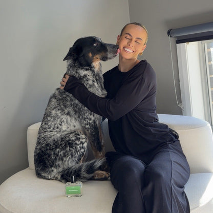 A woman in a black outfit sits on a white sofa hugging a black and white spotted dog, with a dog perfume placed on the sofa in front of her.